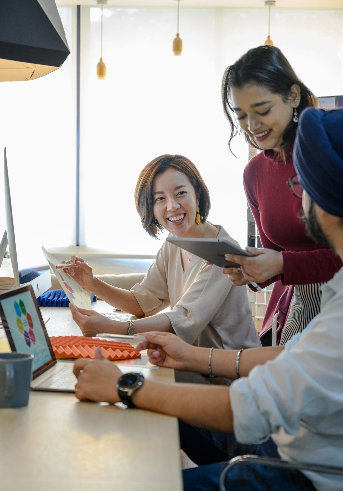 A diverse team laughing together over a tablet and laptop in a bright office